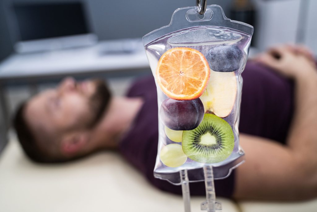 An IV bag filled with colorful fruit hangs beside a man comfortably lying down and receiving Mighty Myers IV Therapy at Revive and Drip. The image highlights the rejuvenating experience provided by this specialized treatment. Revive and Drip offers a wide range of wellness services, including IV Therapy, injectables, Botox, fillers, aesthetic treatments, and personalized weight loss solutions, all designed to enhance your natural beauty and overall well-being.