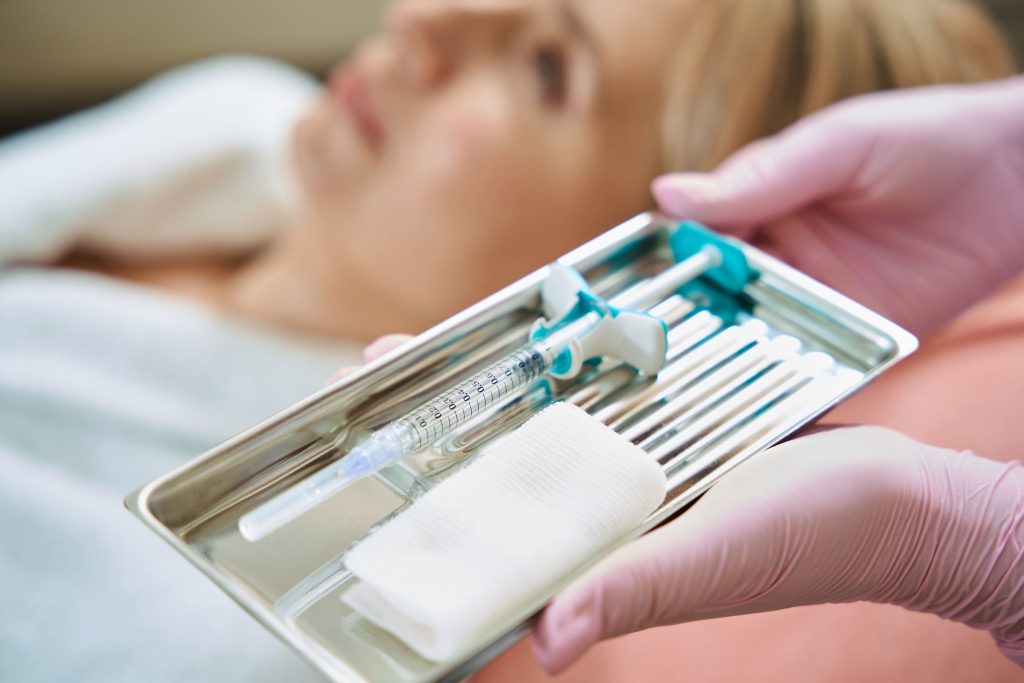 A nurse carefully holding a needle for a Botox injection on a tray, with a woman patiently waiting in the background for her treatment at Revive and Drip. This image highlights the precision and care involved in the process of injections, showcasing the professional and attentive service provided by Revive and Drip. The clinic offers a wide range of aesthetic treatments, including Botox and dermal fillers, as well as IV therapy and weight loss treatments, ensuring that every client receives personalized care to meet their beauty and wellness goals.