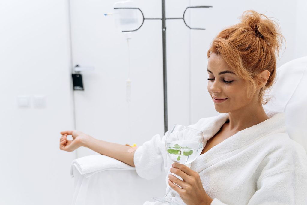 Pretty woman in a white robe, sitting in a recliner with a wide smile and bright white teeth, holding a glass of white wine while receiving IV therapy at Revive and Drip Medical Spa. The spa offers a range of services, including Botox, fillers, aesthetic treatments, IV therapy, and weight loss treatments, helping clients achieve relief from PMS symptoms and enhance their overall well-being in a luxurious and relaxing environment.